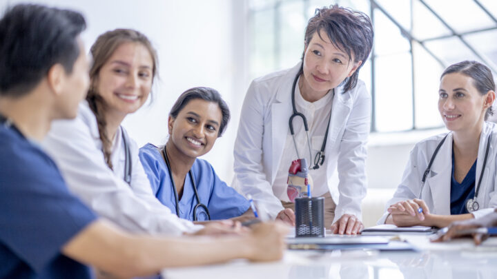 A senior doctor of Asian decent meets with a small group of medical students as she works through their training. She is wearing a white lab coat and has a stethoscope around her neck as she teaches them about the heart.