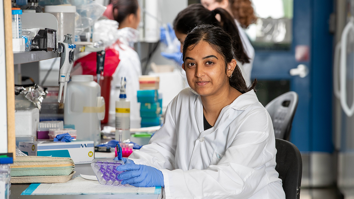 A student is sitting at a lab bench wearing a lab coat and blue gloves. Behind them are other students and lab equipment.