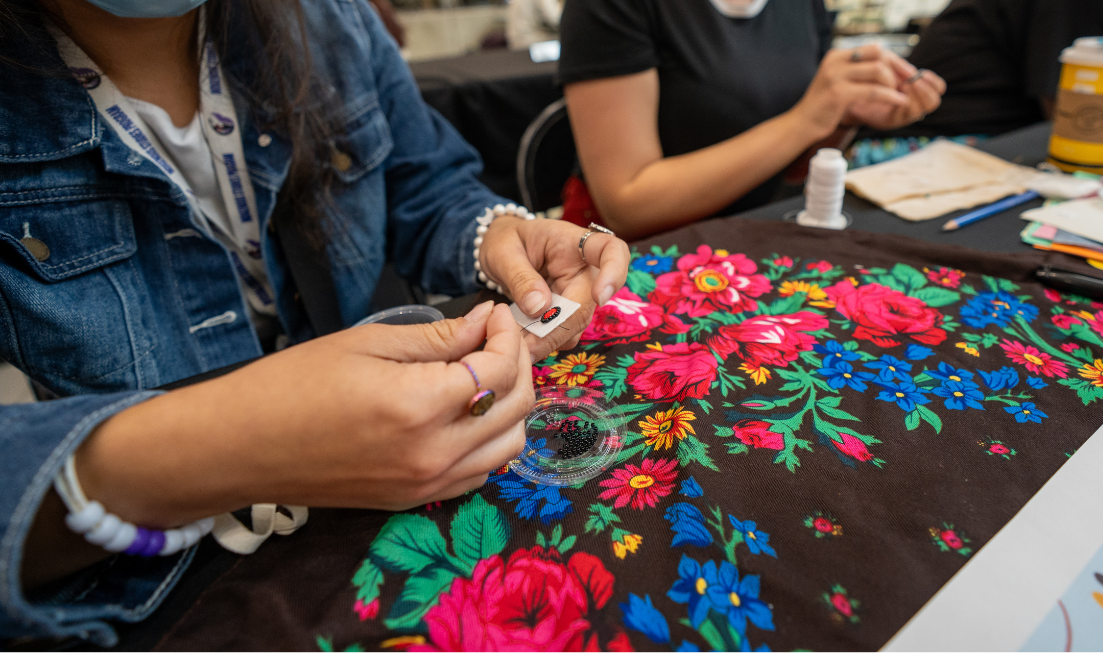 Three women sitting at a table, the one closest to the camera is working on beading.